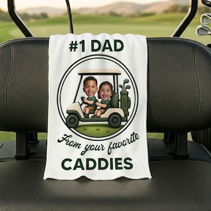 May include: A white waffle-textured golf towel featuring a circular graphic of a golf cart with two children inside. The text reads #1 DAD at the top and From your favorite CADDIES at the bottom, all printed in a dark green color scheme.