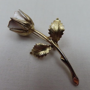 May include: A gold-toned metal rose brooch. The brooch features a detailed rose bud with delicate petals and two leaves. The stem curves gracefully, adding to the realistic design. A classic floral accessory.