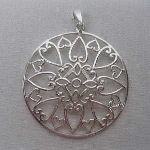 May include: A silver pendant with a circular design. The pendant features an openwork pattern of hearts and swirling lines. The bail is at the top for a chain. The pendant is made of a shiny, reflective metal.