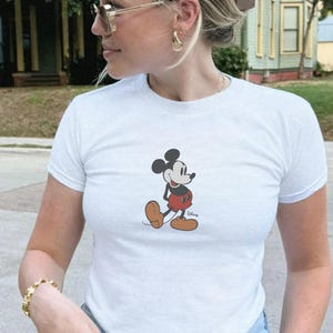 May include: White t-shirt featuring a classic Mickey Mouse graphic. The cartoon character is depicted in his iconic red shorts, black shoes, and gloves. The shirt is short-sleeved and appears to be made of a soft material.
