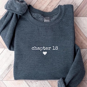 Peut inclure: Sweat-shirt gris foncé avec l'inscription blanche "chapter 18" et un petit cœur blanc. Le sweat-shirt est plié sur une surface en bois.