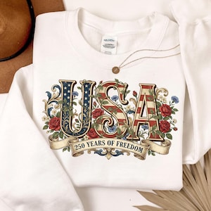 May include: White crew neck sweatshirt featuring a patriotic "USA" design. The letters are decorated with the American flag and floral accents. The text "250 years of freedom" is displayed on a banner below the letters.