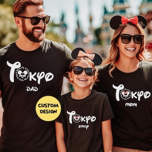 May include: Black t-shirts with a white "Tokyo" design and Mickey Mouse graphic. The shirts are labeled "Dad," "Mom," and "Emily." The family is wearing sunglasses and Mickey Mouse ears. A yellow "Custom Design" sticker is visible.
