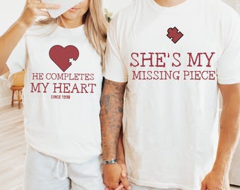 Puede incluir: Dos camisetas blancas a juego con gráficos granate. Una dice "He completes my heart since 1998" con un corazón y una pieza de rompecabezas. La otra dice "She's my missing piece" con una pieza de rompecabezas.