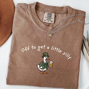 May include: A brown Comfort Colors t-shirt with the phrase "Off to get a little silly" in white. The shirt features a cartoon duck wearing a green hat and jacket, holding a beer mug. A silver necklace with a small pendant is draped on the shirt.