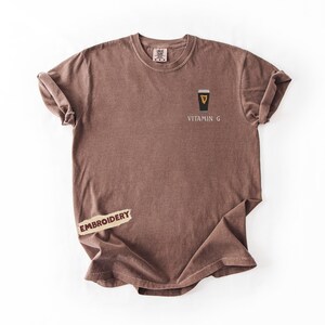 May include: A brown t-shirt with rolled-up sleeves. The shirt features an embroidered design of a black and gold pint glass with the text "VITAMIN G" below it. A small "EMBROIDERY" label is attached to the shirt.