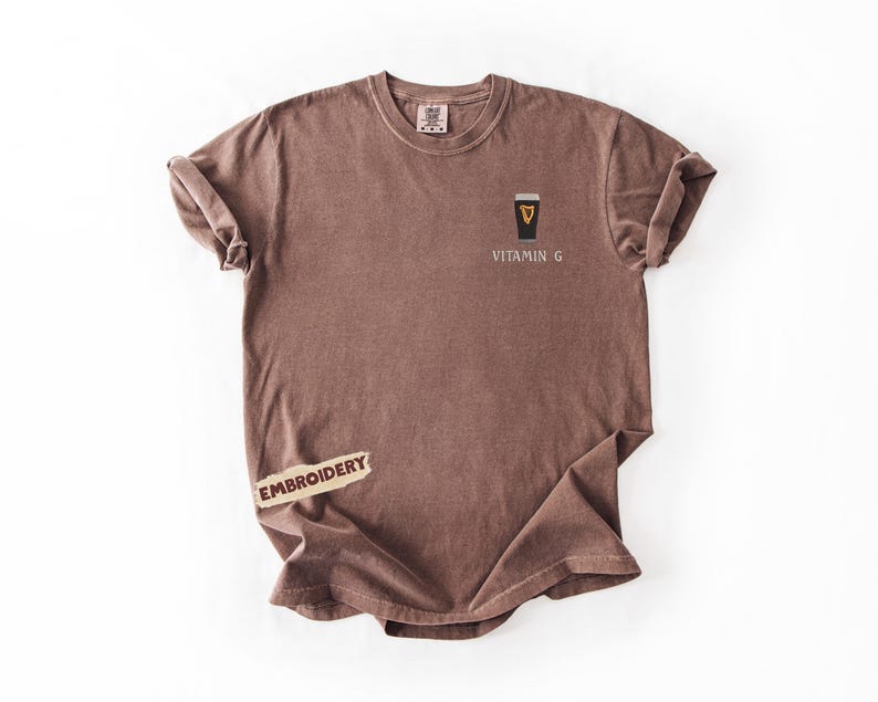 May include: A brown t-shirt with rolled-up sleeves. The shirt features an embroidered design of a black and gold pint glass with the text "VITAMIN G" below it. A small "EMBROIDERY" label is attached to the shirt.