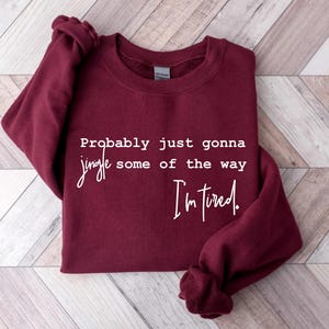 Puede incluir: Una sudadera burdeos con el texto "Probably just gonna jingle some of the way I'm tired." en escritura blanca. La sudadera est&aacute; doblada sobre una superficie clara de vetas de madera.