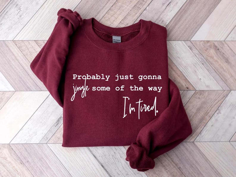 Puede incluir: Una sudadera burdeos con el texto "Probably just gonna jingle some of the way I'm tired." en escritura blanca. La sudadera est&aacute; doblada sobre una superficie clara de vetas de madera.