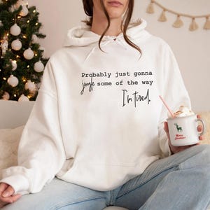 Puede incluir: Sudadera con capucha blanca con el texto "Probably just gonna jingle some of the way, I'm tired." Se sostiene una taza blanca con un reno verde y las palabras "Merry Christmas". Un &aacute;rbol de Navidad est&aacute; en el fondo.