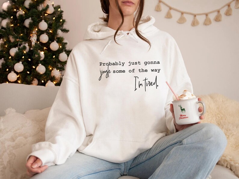 Puede incluir: Sudadera con capucha blanca con el texto "Probably just gonna jingle some of the way, I'm tired." Se sostiene una taza blanca con un reno verde y las palabras "Merry Christmas". Un &aacute;rbol de Navidad est&aacute; en el fondo.
