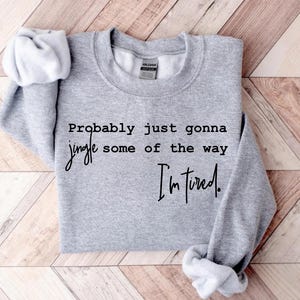 Puede incluir: Una sudadera gris jaspeada con el texto "Probably just gonna jingle some of the way I'm tired." La sudadera tiene cuello redondo y mangas largas. Las mangas est&aacute;n enrolladas. El fondo es un patr&oacute;n de chevron marr&oacute;n claro y blanco.