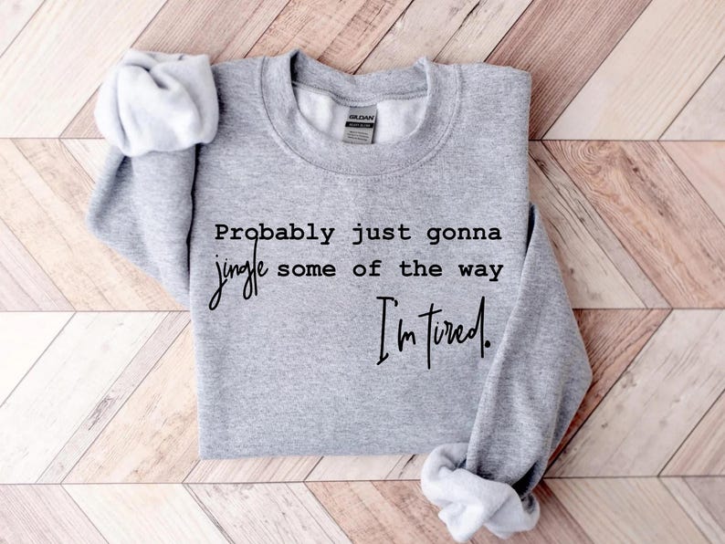 Puede incluir: Una sudadera gris jaspeada con el texto "Probably just gonna jingle some of the way I'm tired." La sudadera tiene cuello redondo y mangas largas. Las mangas est&aacute;n enrolladas. El fondo es un patr&oacute;n de chevron marr&oacute;n claro y blanco.