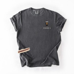 May include: Dark gray t-shirt with rolled-up sleeves. The shirt features an embroidered design of a coffee cup with the text "Vitamin G" below it. A small "Embroidery" tag is attached to the shirt.