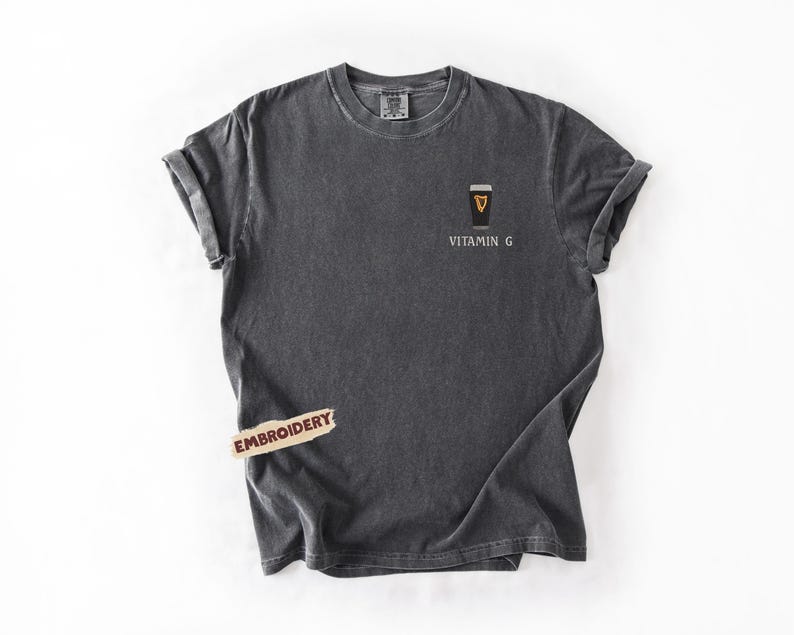 May include: Dark gray t-shirt with rolled-up sleeves. The shirt features an embroidered design of a coffee cup with the text "Vitamin G" below it. A small "Embroidery" tag is attached to the shirt.