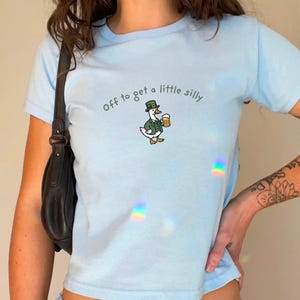 May include: Light blue t-shirt with the phrase "off to get a little silly" and a cartoon duck wearing a green hat and holding a beer mug. The duck is dressed in a green jacket. The shirt is short-sleeved.