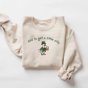 May include: A cream-colored sweatshirt with the phrase "Off to get a little silly" in green script. The graphic features a cartoon duck wearing a green hat and jacket, holding a beer mug.