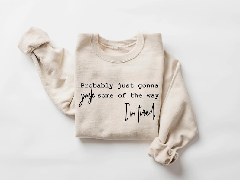 Puede incluir: Sudadera color crema con el texto negro: "Probably just gonna jingle some of the way, I'm tired". La parte superior de cuello redondo y manga larga est&aacute; doblada, con las mangas ca&iacute;das casualmente.