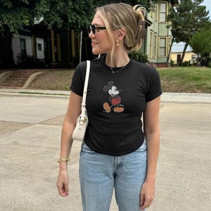 May include: A black t-shirt featuring a Mickey Mouse graphic. The shirt is paired with light blue jeans and a white shoulder bag. The person is wearing sunglasses and gold jewelry.