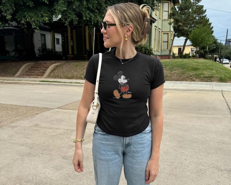 May include: A black t-shirt featuring a Mickey Mouse graphic. The shirt is paired with light blue jeans and a white shoulder bag. The person is wearing sunglasses and gold jewelry.