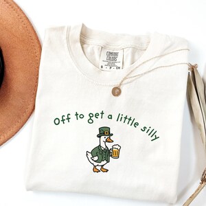 May include: Cream-colored t-shirt with the phrase "Off to get a little silly" in green script. The shirt features a cartoon duck wearing a green jacket and hat, holding a beer stein. A gold necklace is draped across the shirt.