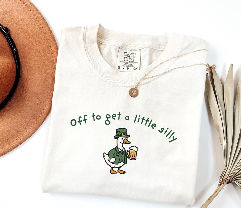 May include: Cream-colored t-shirt with the phrase "Off to get a little silly" in green script. The shirt features a cartoon duck wearing a green jacket and hat, holding a beer stein. A gold necklace is draped across the shirt.