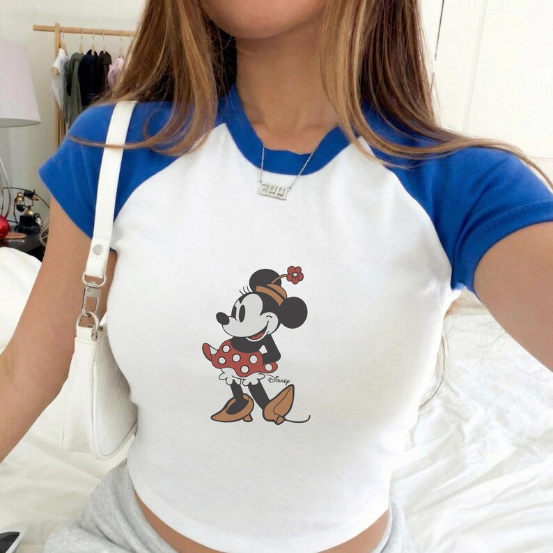 Puede incluir: Camiseta de estilo ragl&aacute;n blanca y azul con un dise&ntilde;o de Minnie Mouse. La camiseta tiene un cuerpo blanco, mangas y cuello azules. El dise&ntilde;o de Minnie Mouse est&aacute; en el centro de la camiseta.