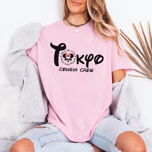 May include: A light pink t-shirt with the word "Tokyo" in black script, featuring a Minnie Mouse graphic and the text "Disney Trip 2024, Family Memories, Cousin Crew". The shirt is a casual style.
