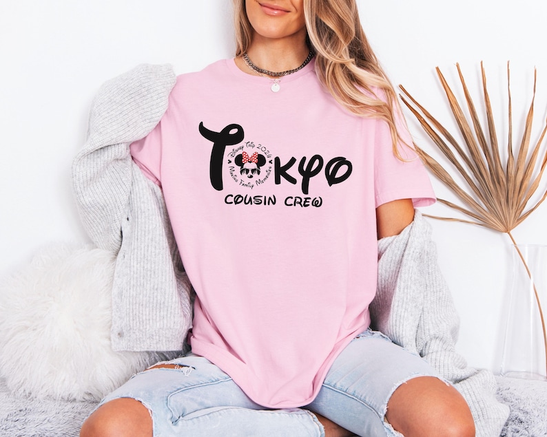 May include: A light pink t-shirt with the word "Tokyo" in black script, featuring a Minnie Mouse graphic and the text "Disney Trip 2024, Family Memories, Cousin Crew". The shirt is a casual style.