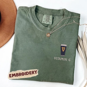 May include: Olive green t-shirt with an embroidered design of a pint of stout and the words "VITAMIN G". A gold necklace and a brown hat are also visible. The word "EMBROIDERY" is printed on a sticker.