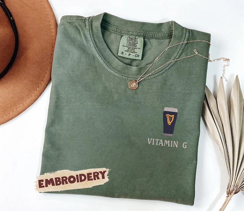 May include: Olive green t-shirt with an embroidered design of a pint of stout and the words "VITAMIN G". A gold necklace and a brown hat are also visible. The word "EMBROIDERY" is printed on a sticker.