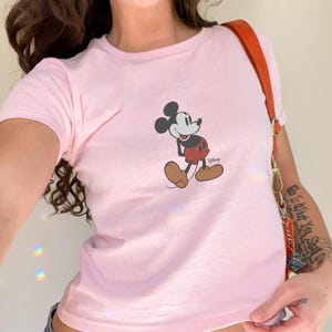 May include: A light pink t-shirt featuring a graphic of Mickey Mouse. The cartoon character is depicted in his classic pose, with a red shorts, brown shoes, and white gloves. The Disney logo is printed below the character.