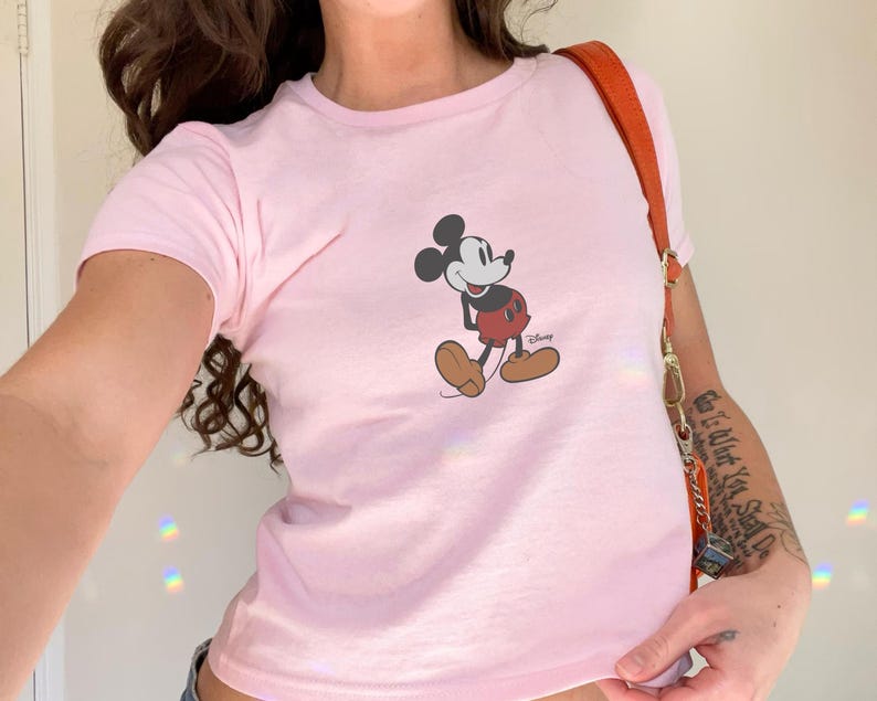 May include: A light pink t-shirt featuring a graphic of Mickey Mouse. The cartoon character is depicted in his classic pose, with a red shorts, brown shoes, and white gloves. The Disney logo is printed below the character.