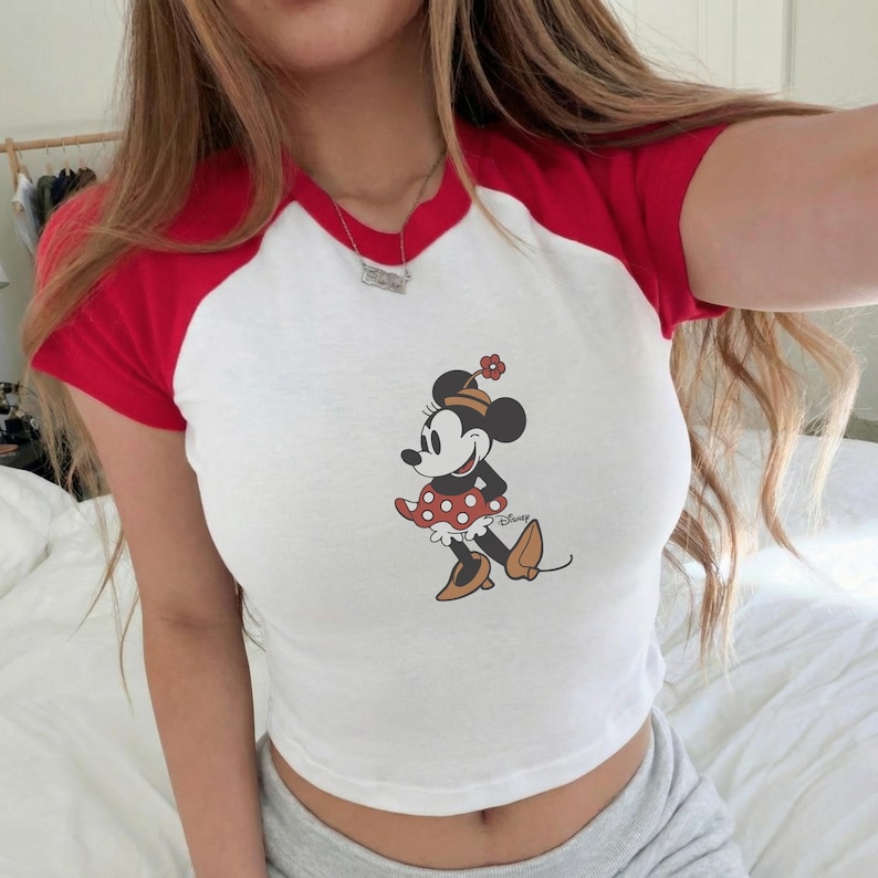 Puede incluir: Top corto blanco con mangas cortas y cuello rojos con un gr&aacute;fico de Minnie Mouse. El personaje de dibujos animados lleva una falda roja de lunares blancos, zapatos marrones y un sombrero con una flor.