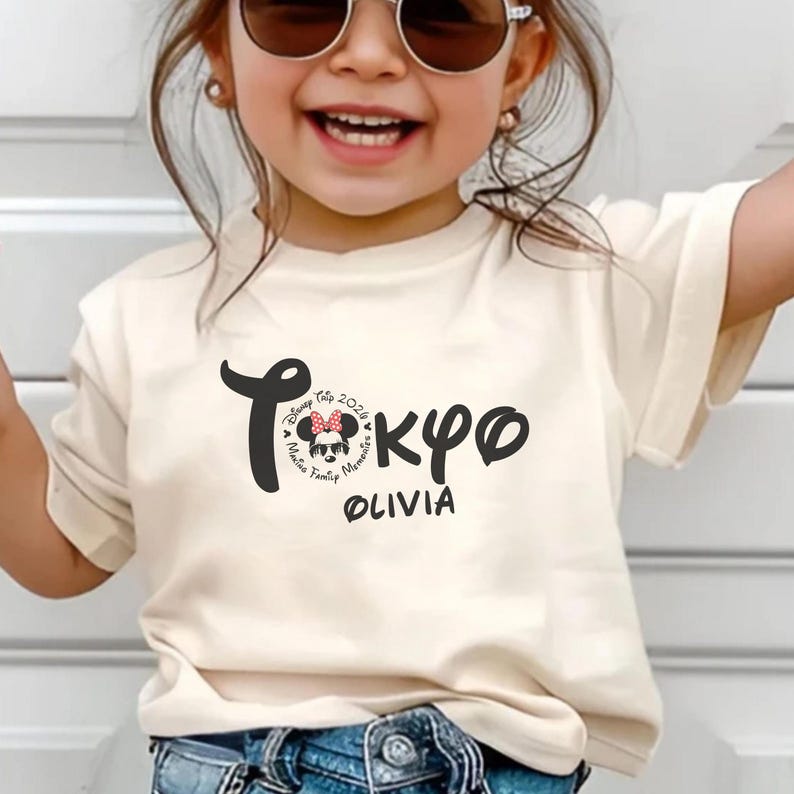 May include: A cream-colored t-shirt featuring the word "Tokyo" in a stylized font, with a Minnie Mouse graphic and the name "Olivia" below. The shirt commemorates a Disney trip in 2024. The child is wearing sunglasses.
