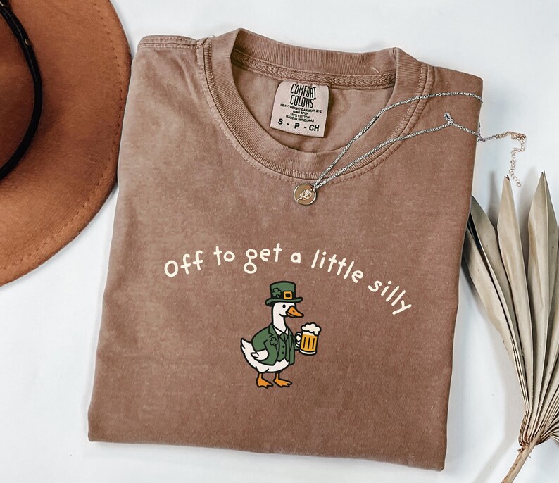 May include: A brown Comfort Colors t-shirt with the phrase "Off to get a little silly" in white. The shirt features a cartoon duck wearing a green hat and jacket, holding a beer mug. A silver necklace with a small pendant is draped on the shirt.