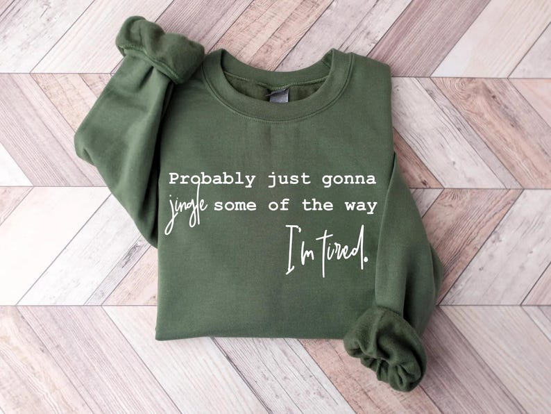 Puede incluir: Una sudadera verde oliva con el texto "Probably just gonna jingle some of the way, I'm tired" en escritura blanca. La sudadera est&aacute; doblada, con las mangas visibles.