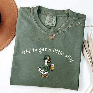 May include: Green t-shirt with the phrase "off to get a little silly" and a cartoon duck wearing a leprechaun hat and holding a beer mug. A gold necklace and a brown hat are also in the image.