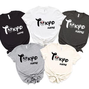 May include: Five short-sleeved t-shirts in black, white, charcoal, gray, and cream. Each shirt features the word "Tokyo" in stylized script, a Mickey Mouse graphic, and the word "NAME" below. The shirts are knotted at the waist.