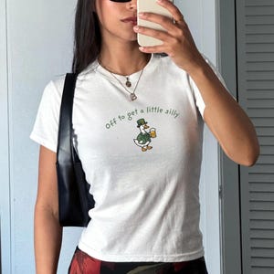 May include: White t-shirt with the text "Off to get a little silly." The shirt features a cartoon duck wearing a green hat and holding a beer mug. The duck is wearing a green jacket and white pants.