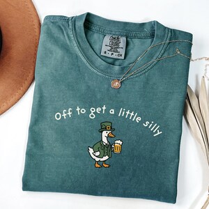 May include: A teal Comfort Colors t-shirt with the phrase "Off to get a little silly" printed in white. The shirt features a cartoon duck wearing a green hat and jacket, holding a beer mug. A gold necklace is draped across the shirt.