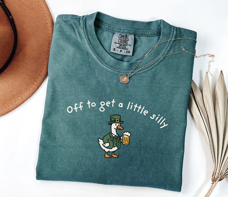 May include: A teal Comfort Colors t-shirt with the phrase "Off to get a little silly" printed in white. The shirt features a cartoon duck wearing a green hat and jacket, holding a beer mug. A gold necklace is draped across the shirt.