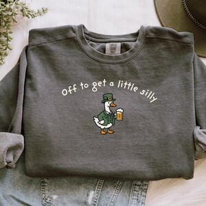 May include: Dark gray sweatshirt with the phrase "off to get a little silly" in white. A cartoon goose wearing a green hat and jacket holds a beer mug. The sweatshirt is folded on top of denim.