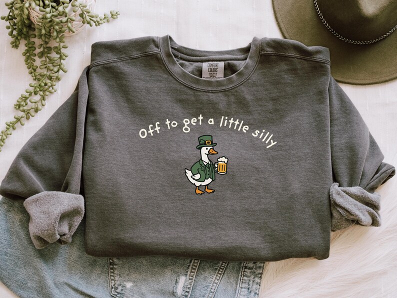 May include: Dark gray sweatshirt with the phrase "off to get a little silly" in white. A cartoon goose wearing a green hat and jacket holds a beer mug. The sweatshirt is folded on top of denim.