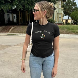 May include: Black t-shirt with the phrase "Off to get a little silly" and a cartoon duck wearing a green hat and holding a beer. The person is wearing sunglasses, gold jewelry, and light blue jeans.