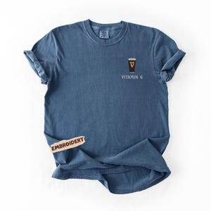 May include: A blue t-shirt with a small embroidered design of a pint of stout with the text "Vitamin G". The shirt has rolled-up sleeves and a tag that says "Embroidery".