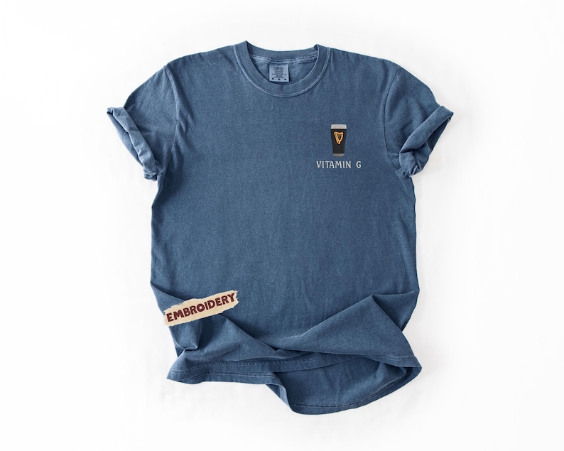 May include: A blue t-shirt with a small embroidered design of a pint of stout with the text "Vitamin G". The shirt has rolled-up sleeves and a tag that says "Embroidery".
