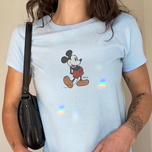 May include: Light blue t-shirt featuring a graphic of Mickey Mouse. The cartoon character is depicted in his classic outfit with red shorts, yellow shoes, and white gloves. The shirt has short sleeves and a crew neck.