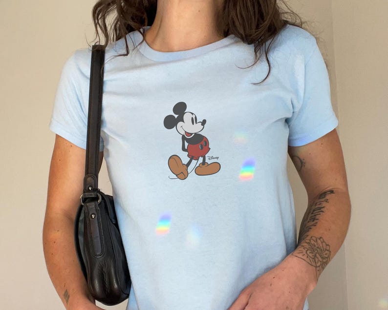 May include: Light blue t-shirt featuring a graphic of Mickey Mouse. The cartoon character is depicted in his classic outfit with red shorts, yellow shoes, and white gloves. The shirt has short sleeves and a crew neck.
