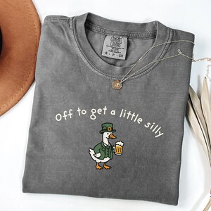 May include: A gray Comfort Colors t-shirt with the phrase "Off to get a little silly" in white. The shirt features a cartoon duck wearing a green hat and jacket, holding a beer. A gold necklace is draped across the shirt.